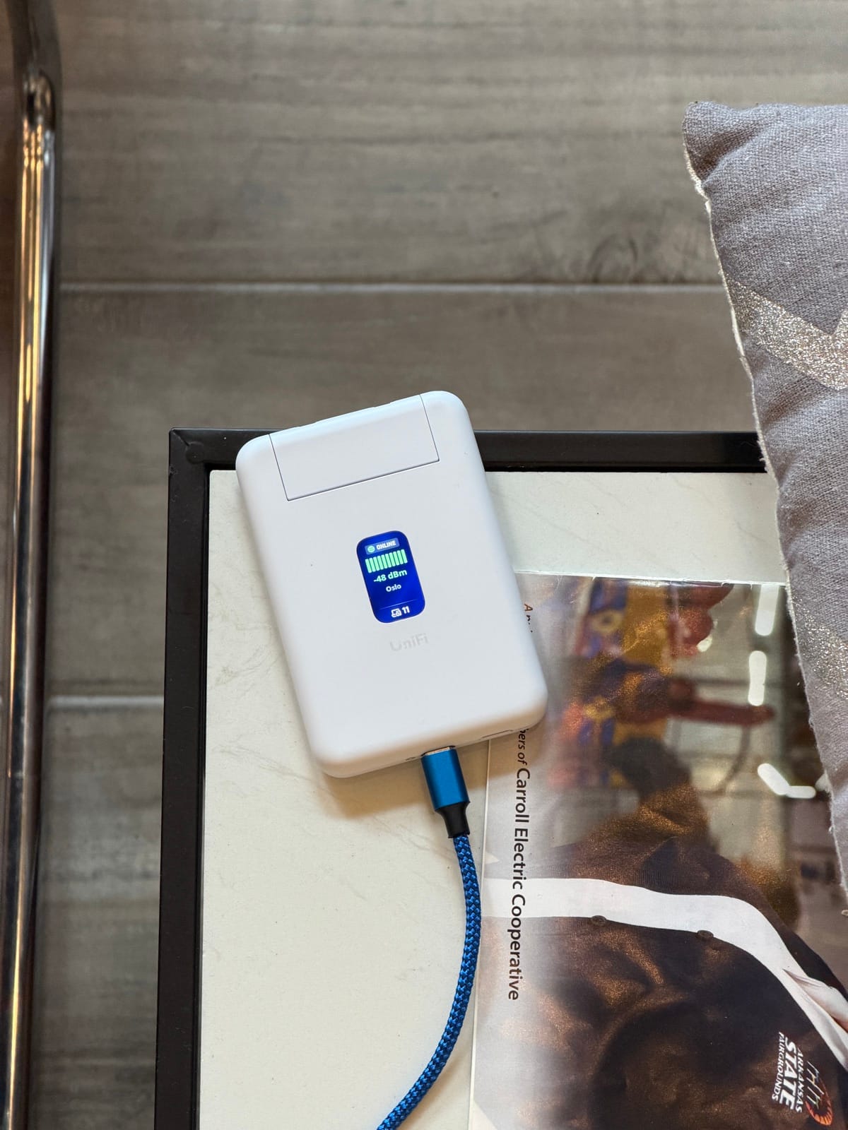 A white UniFi Travel Router on a side table, status screen lit, a braided USB-C cable plugged into its side.