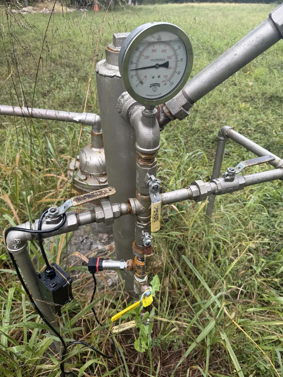 The wellhead: a tall knockout chamber with a mechanical pressure gauge on a side leg, the stainless transmitter threaded into a brass tee below the gauge between two ball valves, and the black sensor node strapped to a pipe at lower left with its cable running to the transmitter