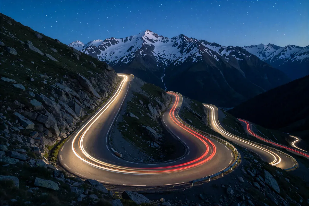Long-exposure light trails climbing a mountain switchback road at dusk under snowy peaks