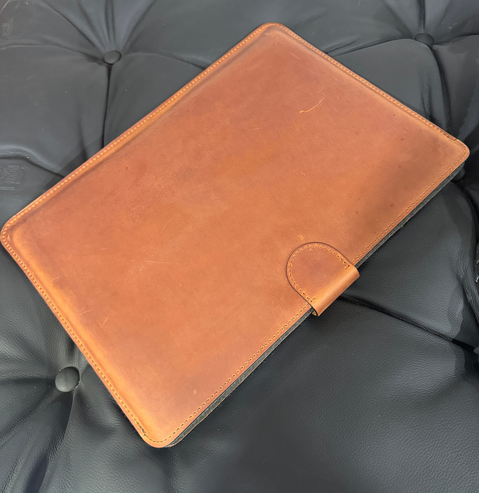 Closed veg-tanned leather MacBook sleeve on a black tufted leather couch, cognac patina visible