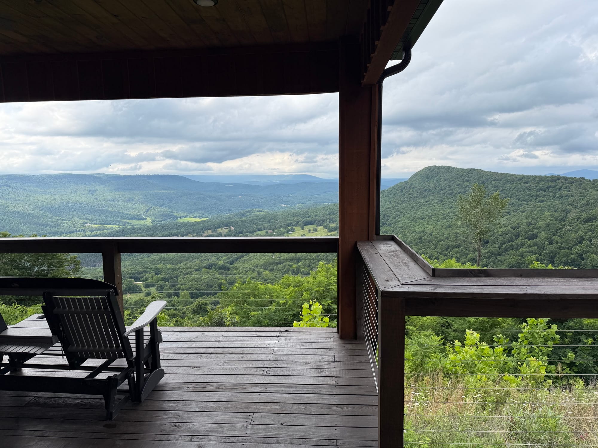 A covered cabin porch with wooden chairs overlooking a wide green mountain valley under an overcast sky