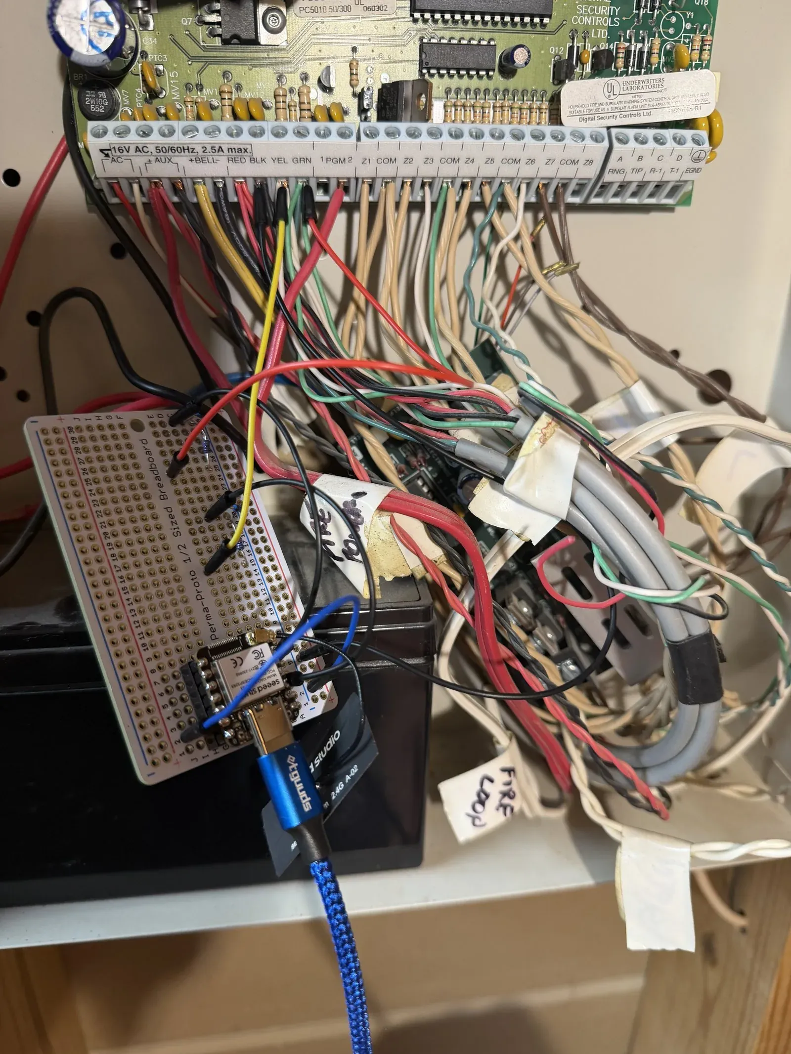 A XIAO ESP32-S3 on a proto board with its voltage-divider wires landed on the alarm panel's labeled screw-terminal strip